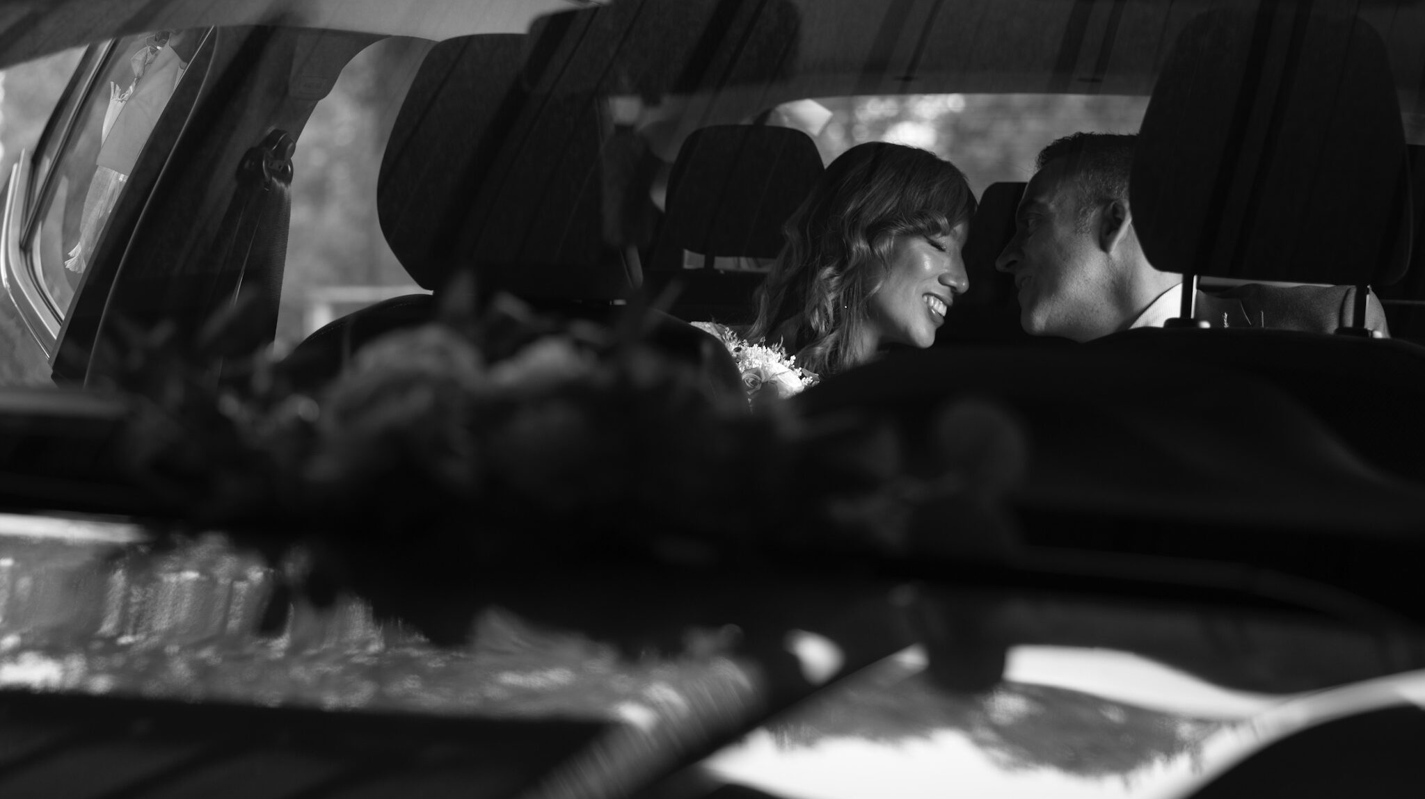 Los novios riendo frente a frente dentro del coche, ramo desenfocado en primer plano