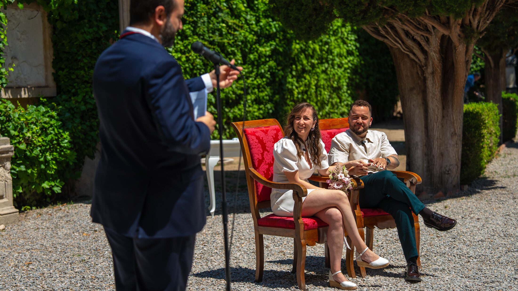 Los novios sentados durante la ceremonia, escuchando al oficiante en un jardín