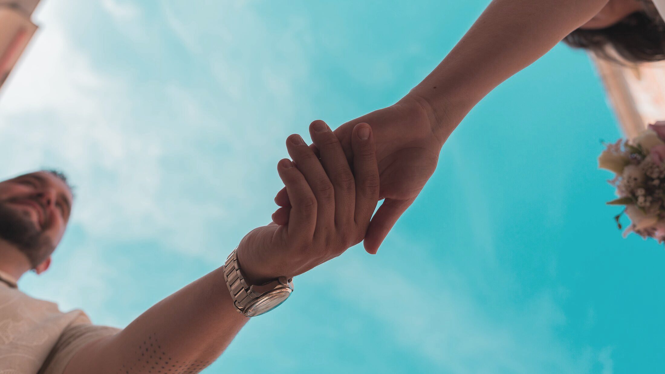 Manos entrelazadas de los novios contra un cielo turquesa, vistas desde abajo