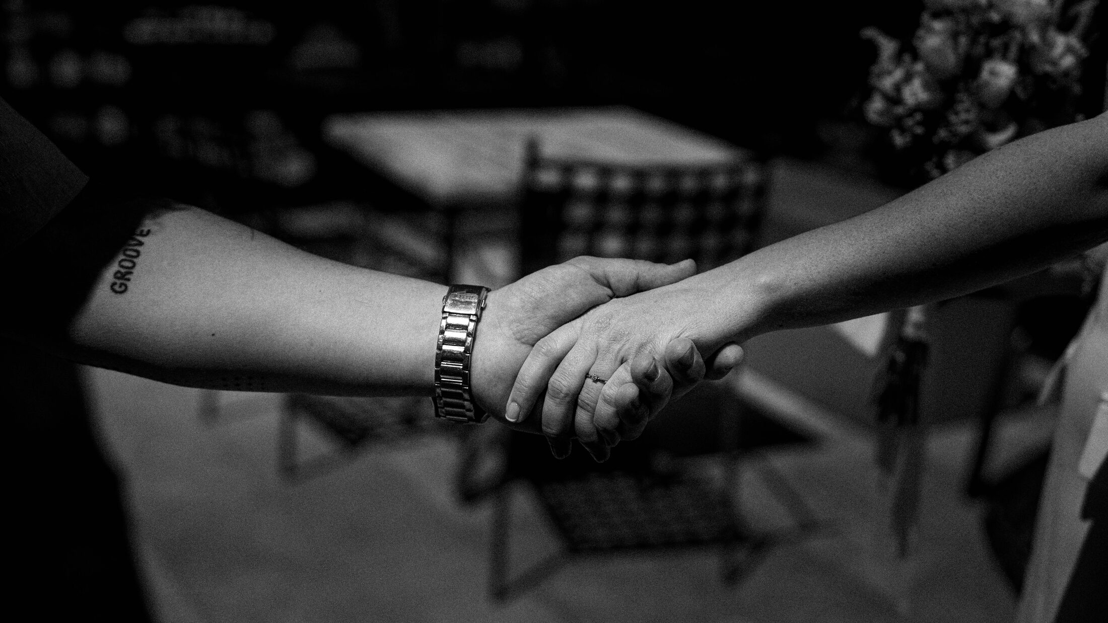 Manos entrelazadas en blanco y negro, con un ramo desenfocado al fondo