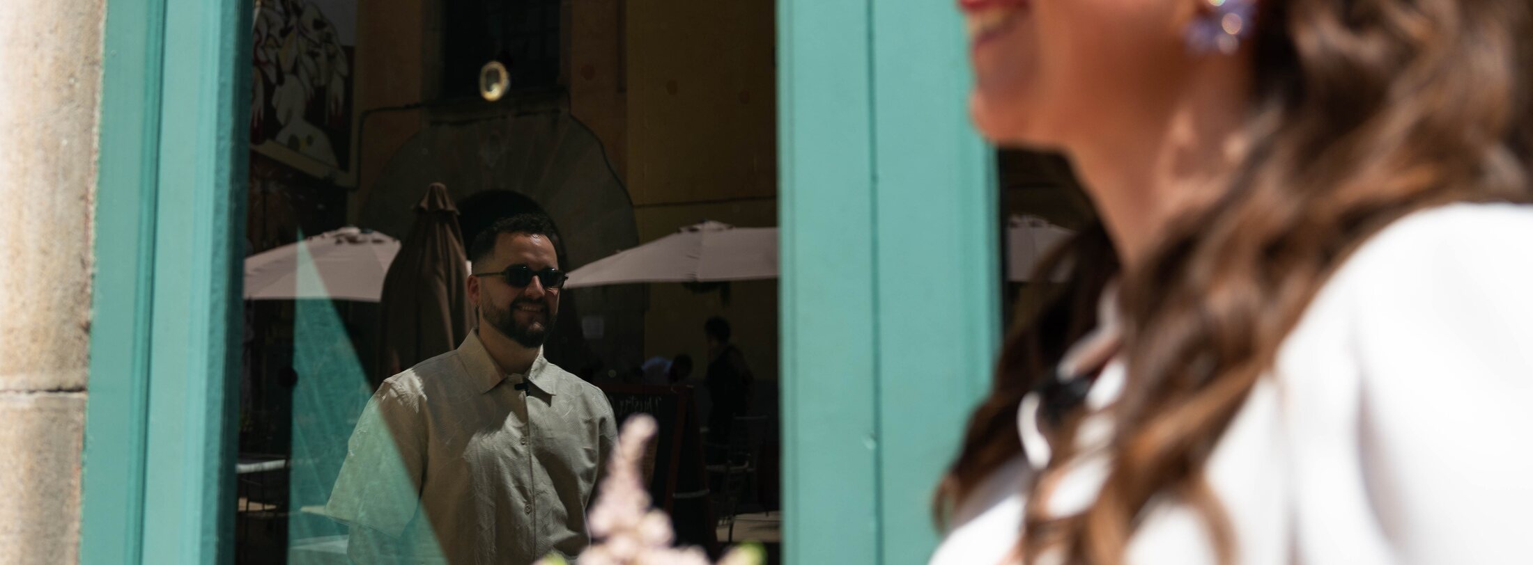 Reflejo del novio sonriendo en el cristal de una puerta turquesa, mientras la novia pasa desenfocada en primer plano